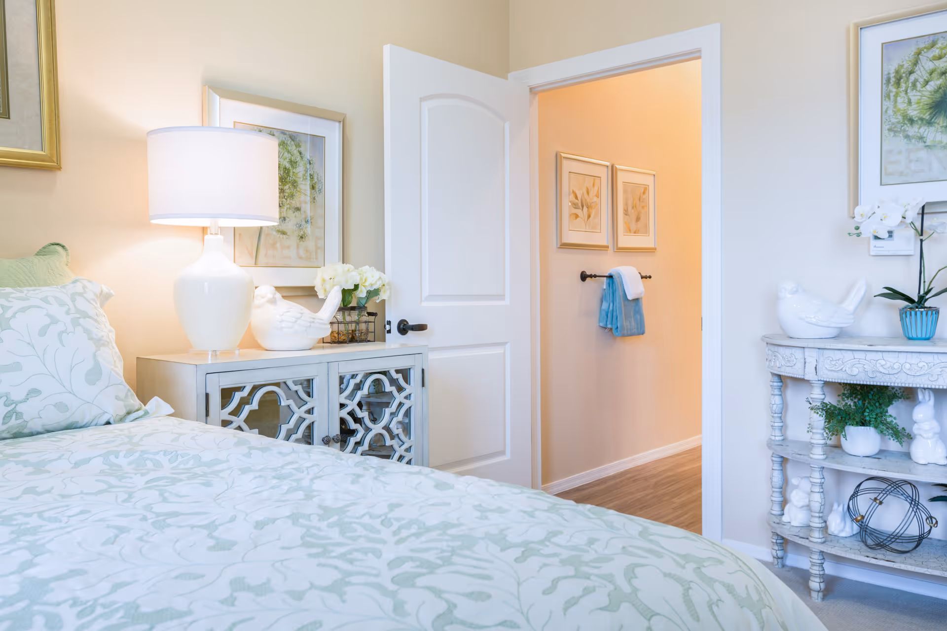 A cozy bedroom with a bed covered in a light green and white patterned bedspread. Next to the bed is a white nightstand with a white lamp, a decorative bird figurine, and a small vase of flowers. The door to the room is open, revealing a hallway with two framed pictures and a towel rack holding blue and white towels. On the right side of the image, there is a decorative white console table with plants and more bird figurines.