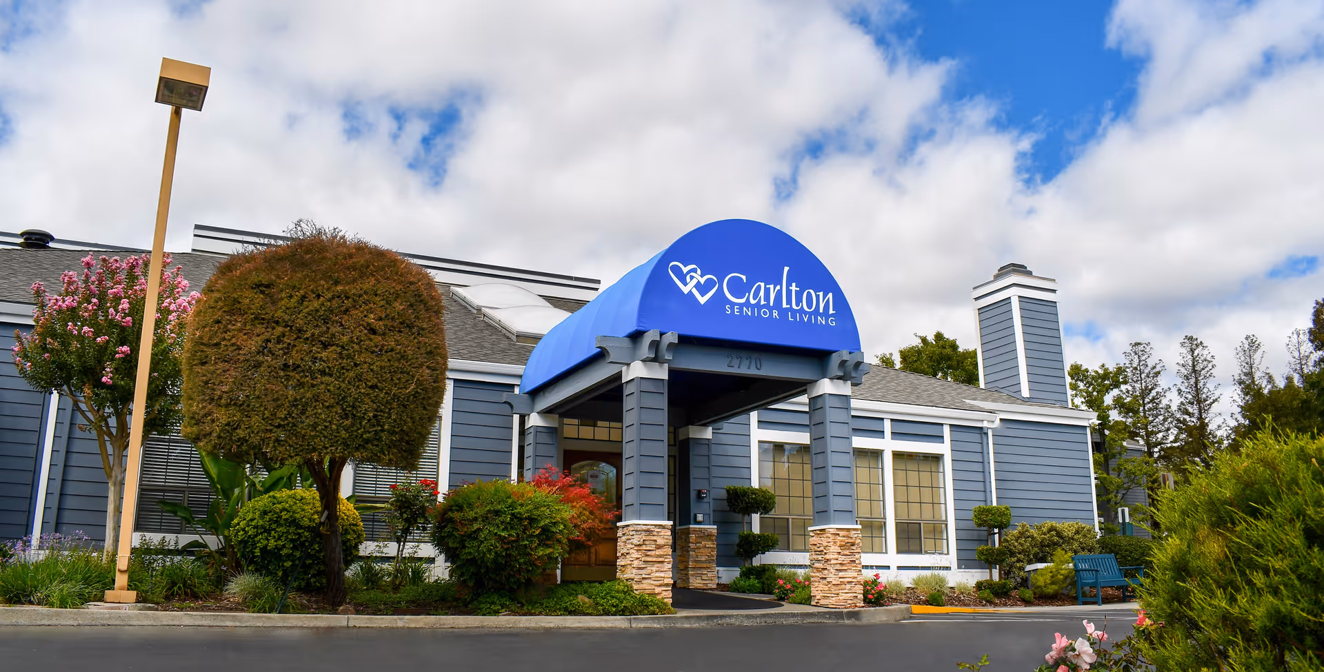 Exterior view of Carlton Senior Living Pleasant Hill - Martinez building with a blue awning over the entrance displaying the facility name. The building is gray with white trim, surrounded by neatly trimmed bushes, flowering plants, and a clear sky with some clouds.