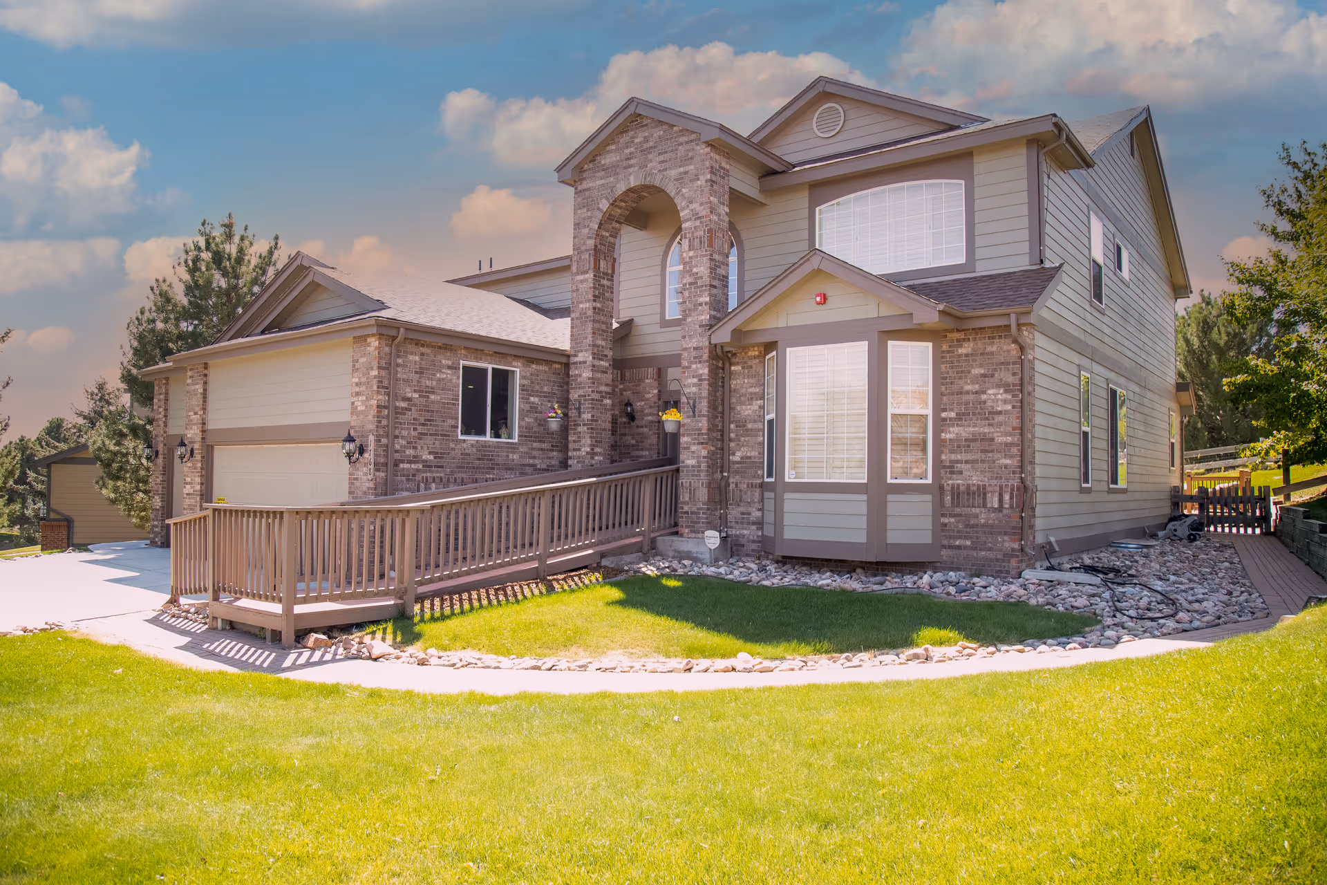 Exterior view of a two-story assisted living facility building with brick and siding facade, a wheelchair accessible ramp, green lawn, and a partly cloudy sky.