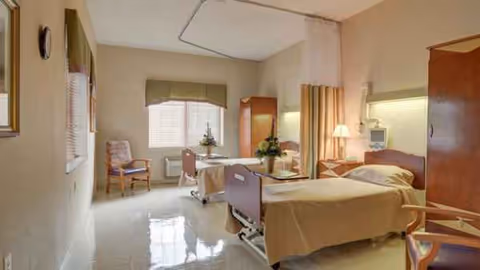 Interior of a senior facility patient room with two beds, chairs, and a window.