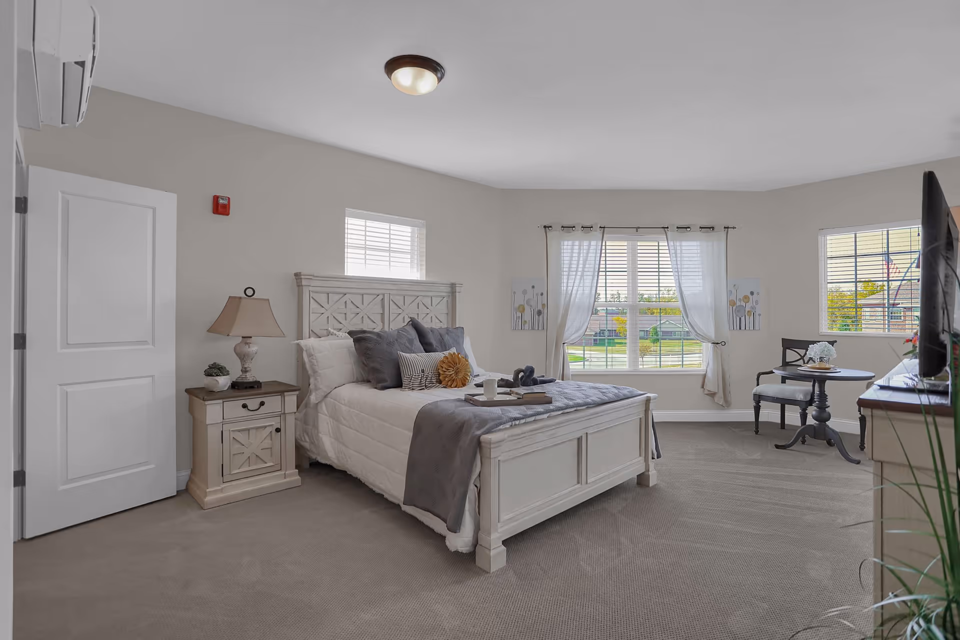 A bright and spacious bedroom with a large bed featuring white and gray bedding and decorative pillows. There is a nightstand with a lamp and a small plant next to the bed. The room has beige walls, carpeted floor, and multiple windows with white blinds and sheer curtains letting in natural light. A small round table with two chairs is placed near the windows, and a flat-screen TV is visible on a dresser to the right.