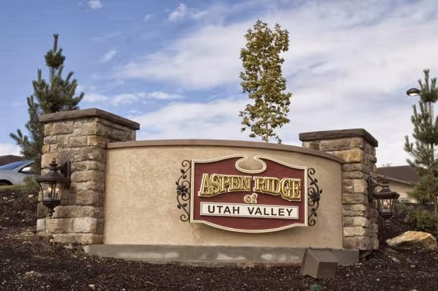Stone and stucco sign for Aspen Ridge of Utah Valley with decorative lanterns on either side, set against a partly cloudy sky and surrounded by small trees and landscaping.