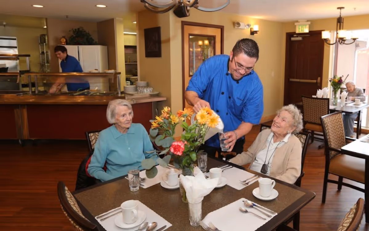 A staff member in a blue shirt pours water for two elderly women seated at a dining table with a vase of flowers in an assisted living dining room.