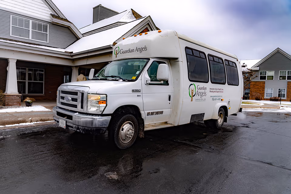 A white Guardian Angels shuttle van parked outside a senior living facility building on a wet driveway with some snow on the ground and roof. The building has a covered entrance with white columns and large windows.