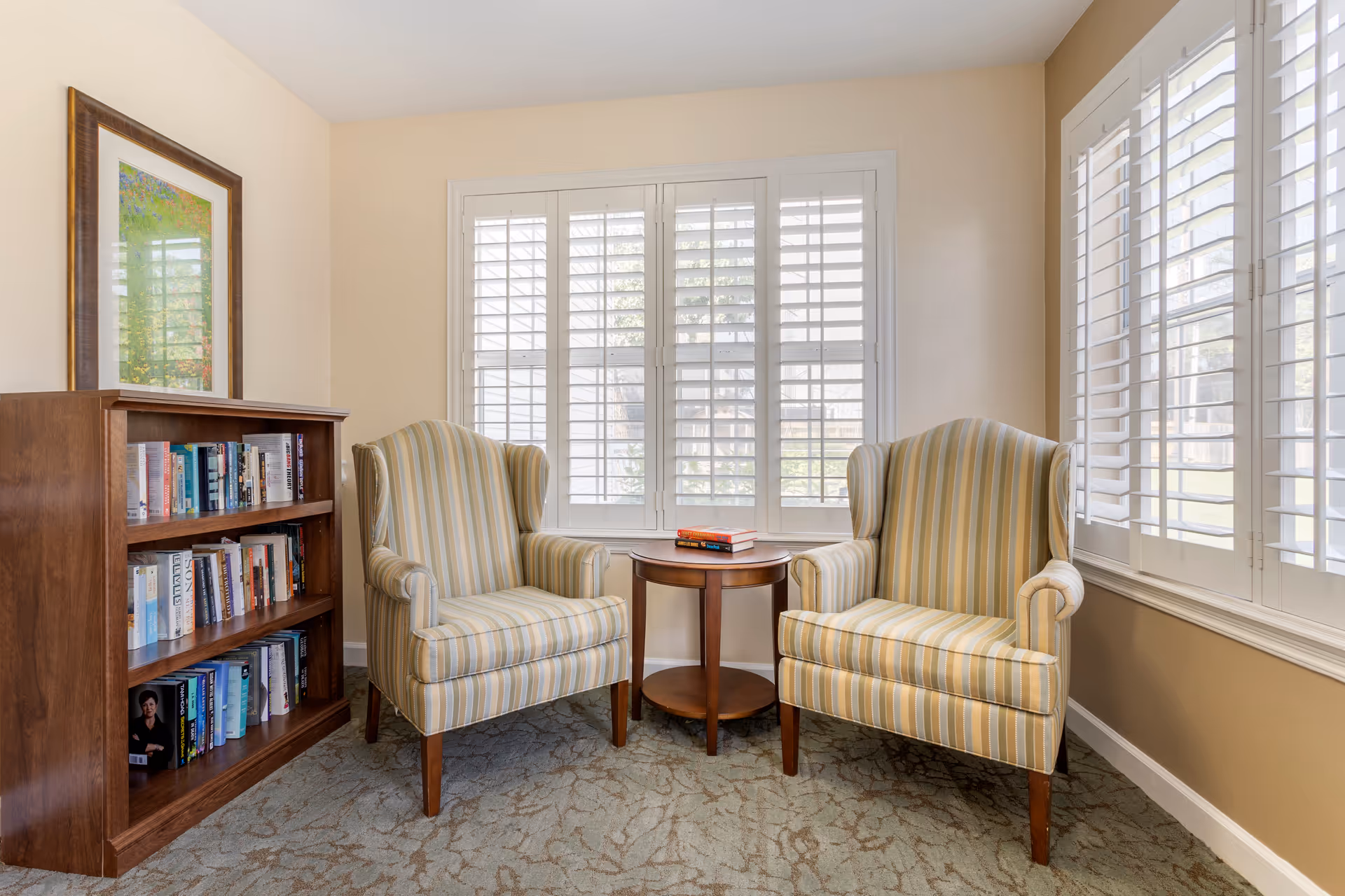 A cozy sitting area with two striped armchairs facing each other with a small round wooden table between them holding three books. To the left is a wooden bookshelf filled with books and a framed artwork hanging above it. The room has beige walls and large windows with white plantation shutters letting in natural light.
