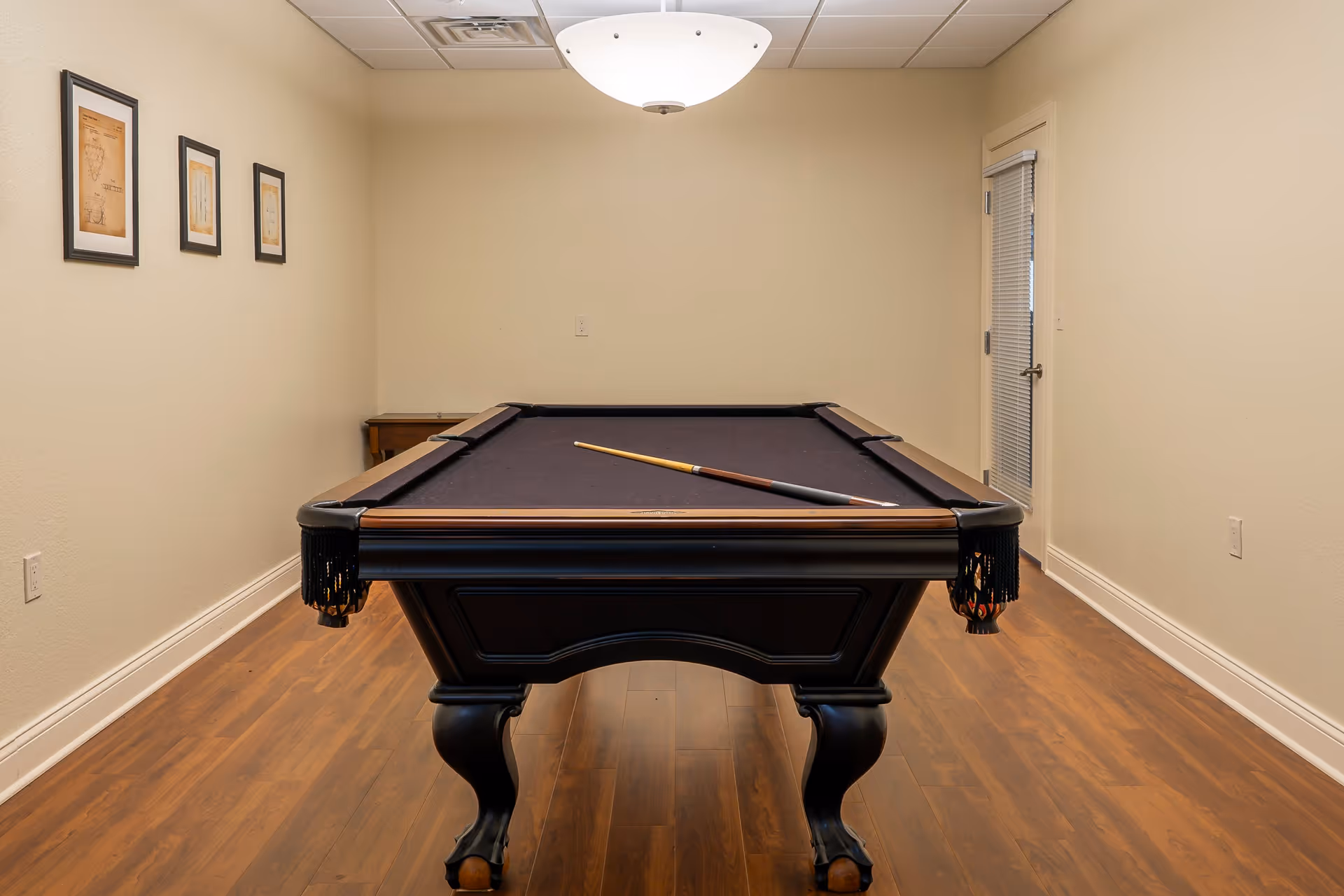 A room with a black pool table centered on a wooden floor. A pool cue rests on the table. The walls are beige with three framed pictures on the left wall. A white ceiling light fixture hangs above the pool table. There is a door with blinds on the right side of the room.