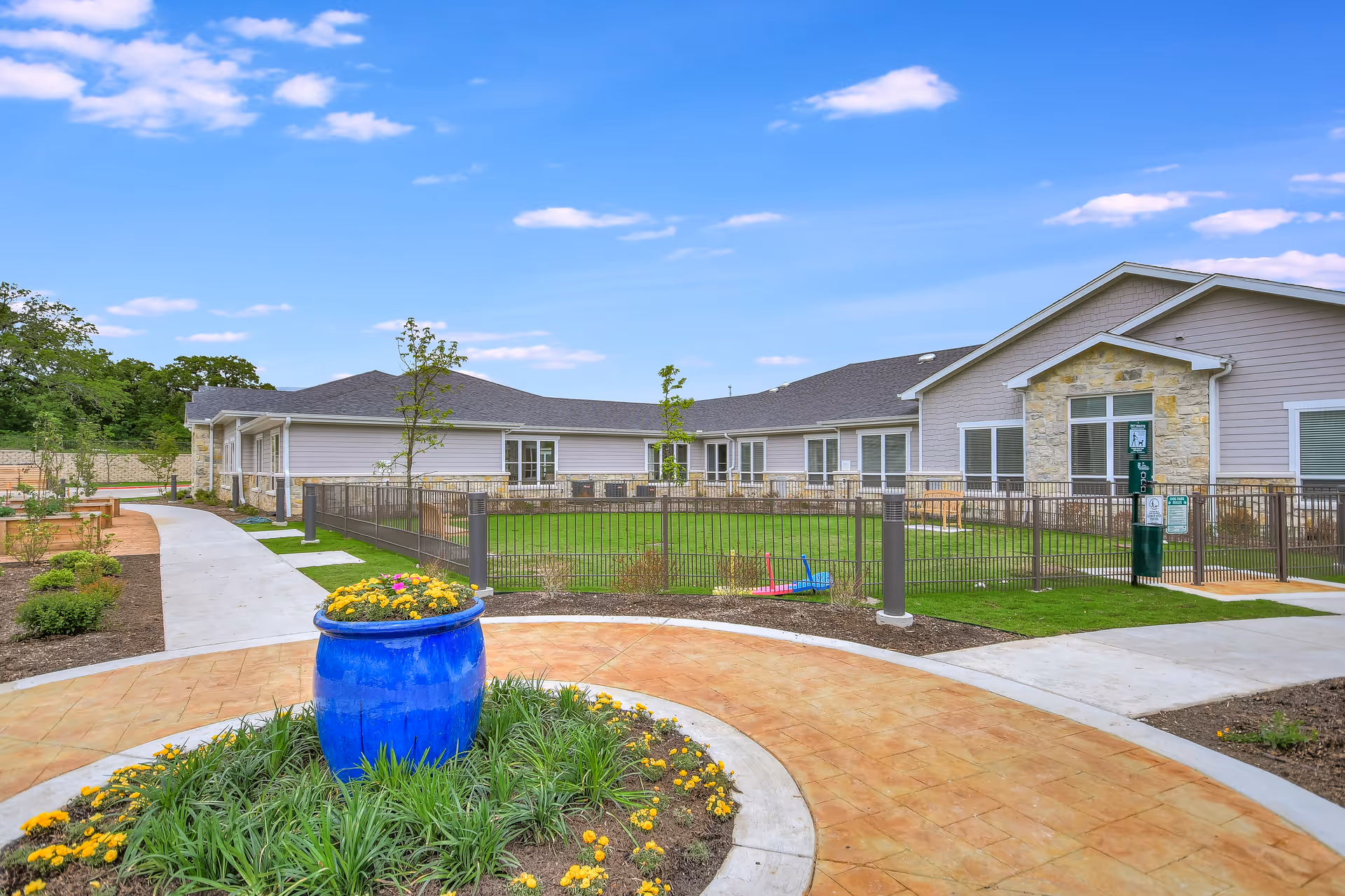 Outdoor view of Legacy Oaks of Azle Senior Living facility showing a landscaped garden area with a large blue flower pot filled with yellow flowers in the foreground, paved walkways, green grass enclosed by a metal fence, and a single-story building with stone and siding exterior under a blue sky with scattered clouds.