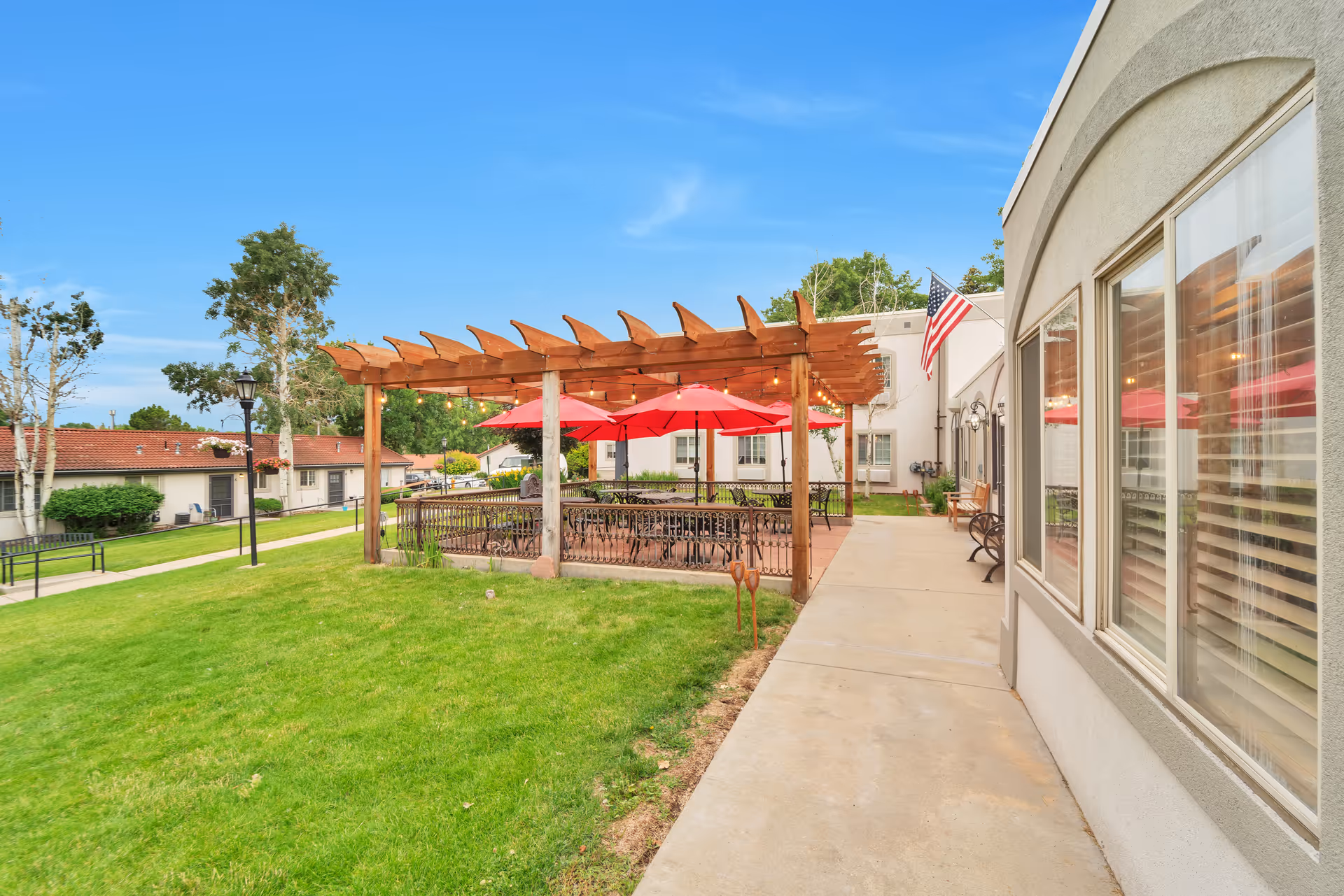 Outdoor patio area at Gardens Care Senior Living - Homestead featuring a wooden pergola with string lights, several red umbrellas shading tables and chairs, a concrete walkway, green grass lawn, benches, and an American flag mounted on the building.