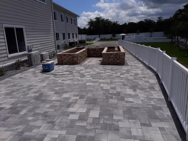 Paved outdoor patio with raised brick planters next to a white fence and a multi-story building under a partly cloudy sky.