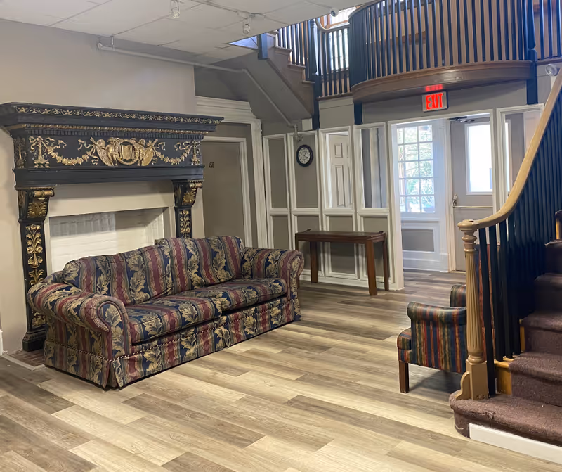 Sitting area with a patterned sofa and armchair in front of an ornate fireplace and a staircase leading to an upper balcony.