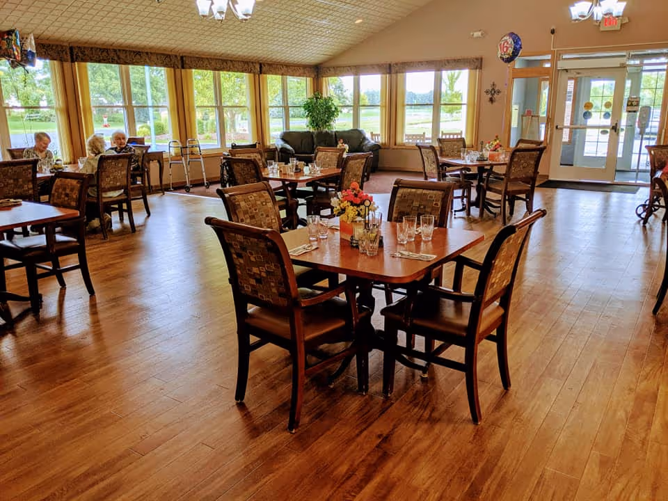 A bright dining room with wooden floors and multiple tables set with glasses and utensils. Several elderly people are seated at a table near large windows that let in natural light. There is a sofa and a walker near the windows, and a door leading outside is visible in the background.