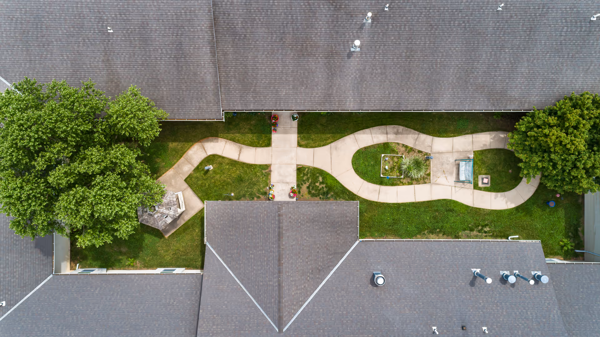 Aerial view of a winding concrete pathway through a green lawn area between buildings with gray shingled roofs. The pathway features a small gazebo on the left side and a bench near a fire pit on the right side, surrounded by trees and landscaping.