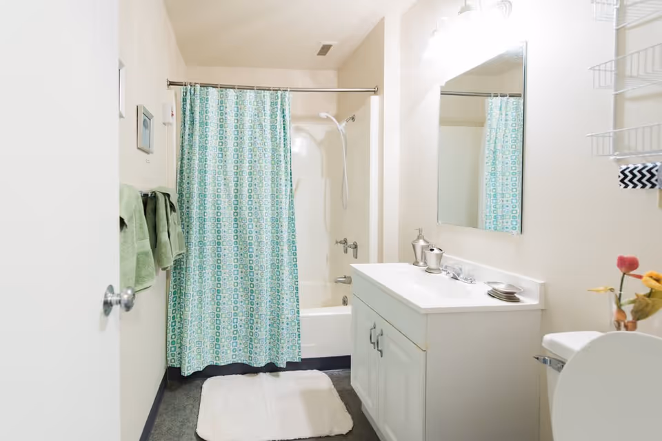 A clean bathroom with a white bathtub and shower combination featuring a blue and green patterned shower curtain. There is a white vanity with a sink, silver soap dispenser, and a mirror above it. Green towels hang on a rack on the left wall, and a white bath mat is placed on the floor in front of the tub. A small vase with flowers is on the right side near the toilet.