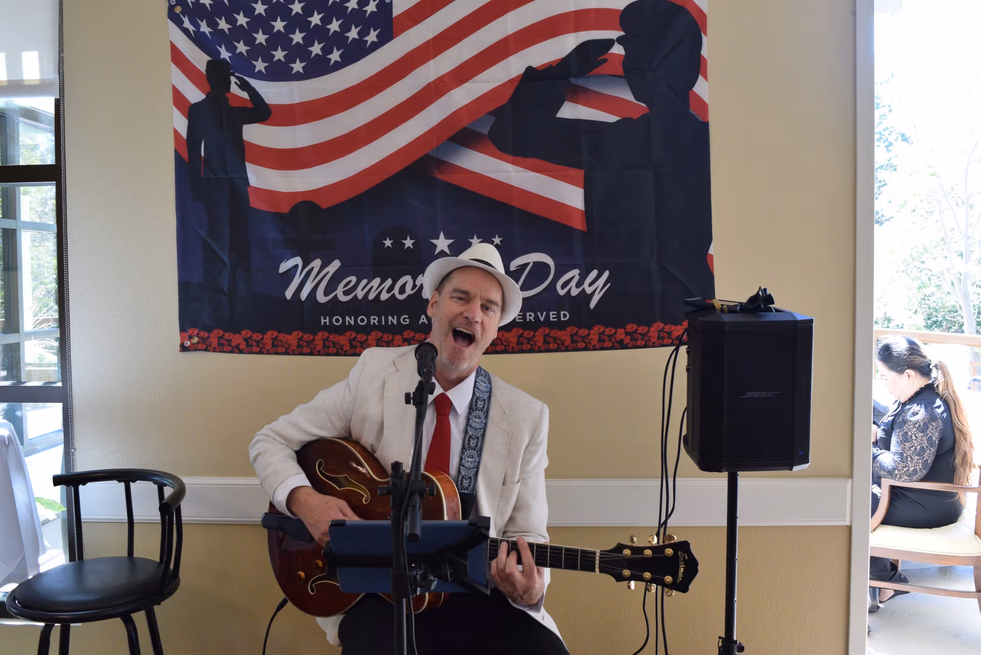 A man wearing a white hat, white jacket, and red tie is playing an acoustic guitar and singing into a microphone. Behind him is a Memorial Day banner with an American flag and silhouettes of saluting soldiers. To the right, a woman is seated near a window, looking down.
