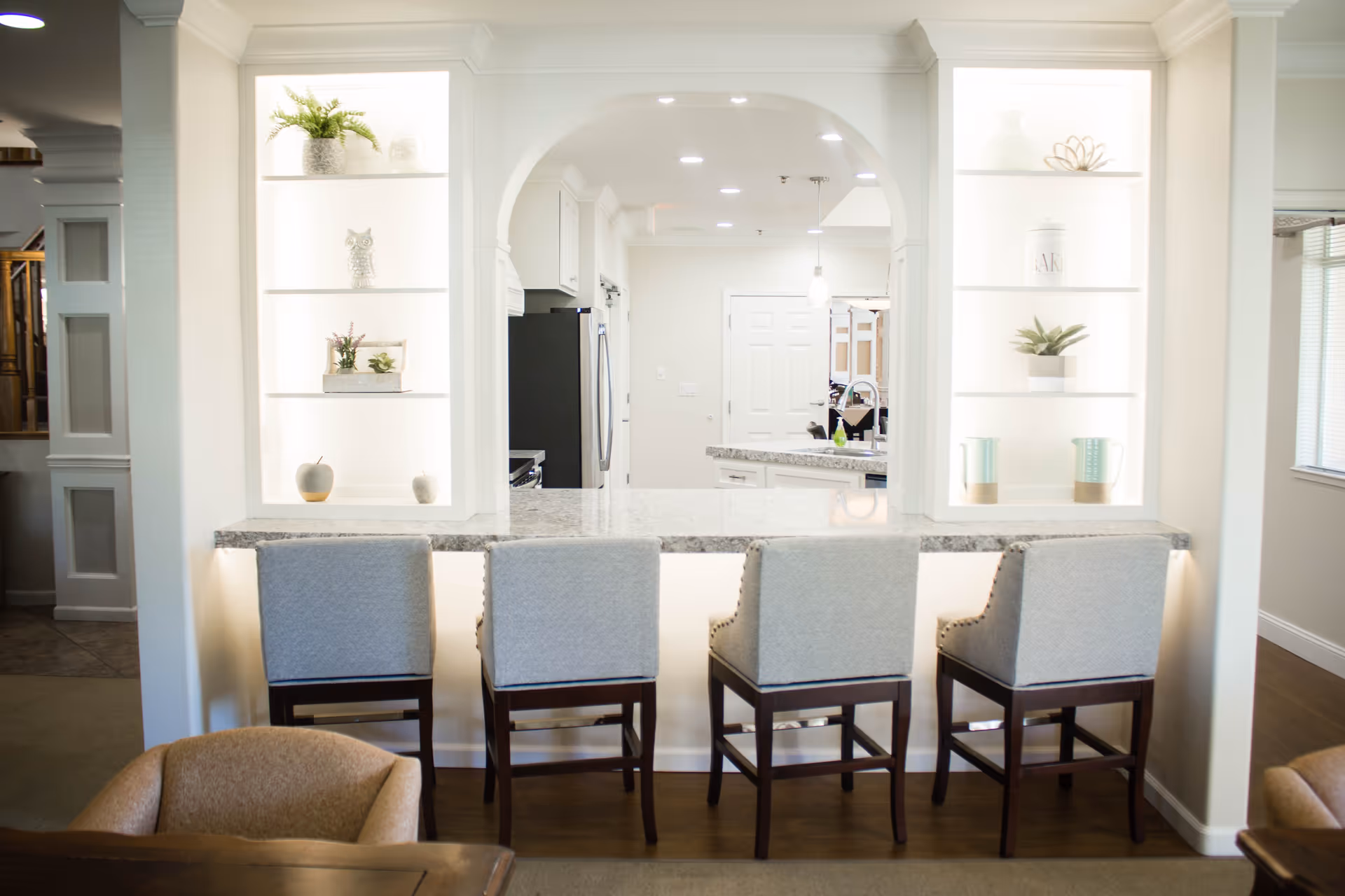 Bar-height marble counter with four upholstered stools facing an open kitchen pass-through flanked by lit display shelves.