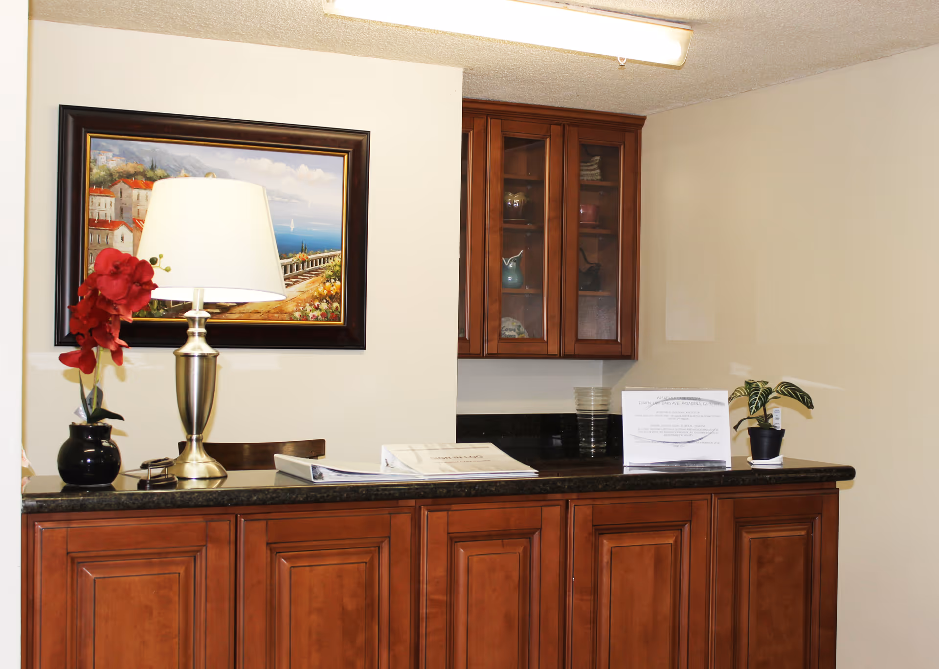 Reception desk area with a dark granite countertop and wooden cabinets below and above. On the counter, there is a silver lamp with a white shade, a small black vase with red flowers, some papers, and a small potted plant. A framed painting of a coastal scene hangs on the wall behind the desk.