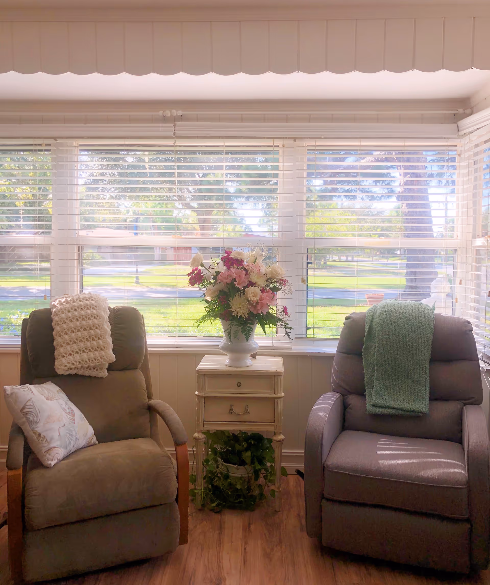 Two recliner chairs flank a small side table topped with a vase of flowers in front of a large window with blinds showing a sunlit lawn outside.