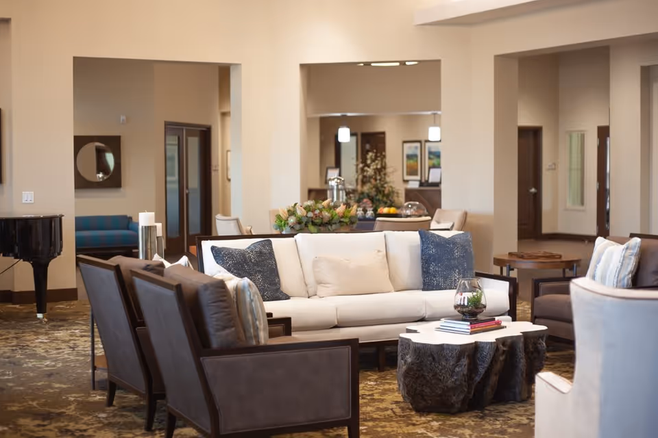 A cozy living room area in an assisted living facility featuring a white sofa with blue and beige pillows, surrounded by several dark brown armchairs. A rustic wooden coffee table with books and a glass terrarium sits in the center. In the background, there is a black grand piano, a blue bench, and a table with decorative plants and fruit. The room has beige walls and carpeted flooring.