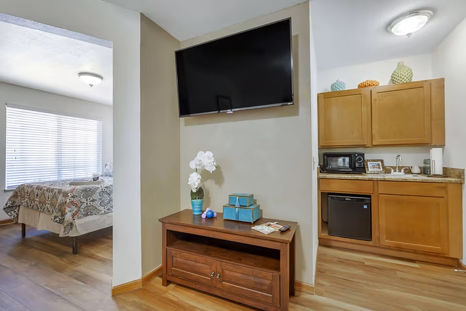 Studio-style apartment interior with a wall-mounted TV above a wooden console, a small kitchenette with cabinets and mini-fridge, and a bed visible through an open doorway.