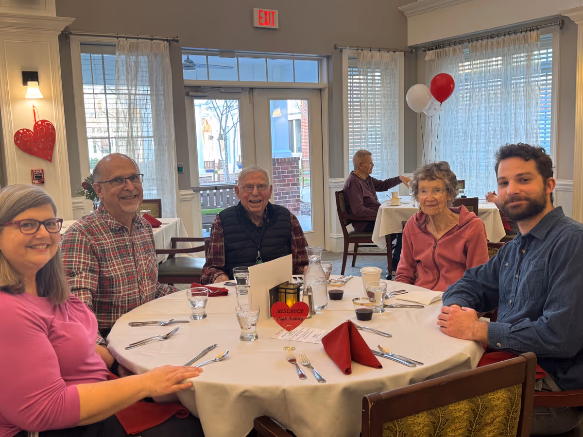 Several people sit around a round table set for a meal in a bright dining room with balloons and heart decorations.