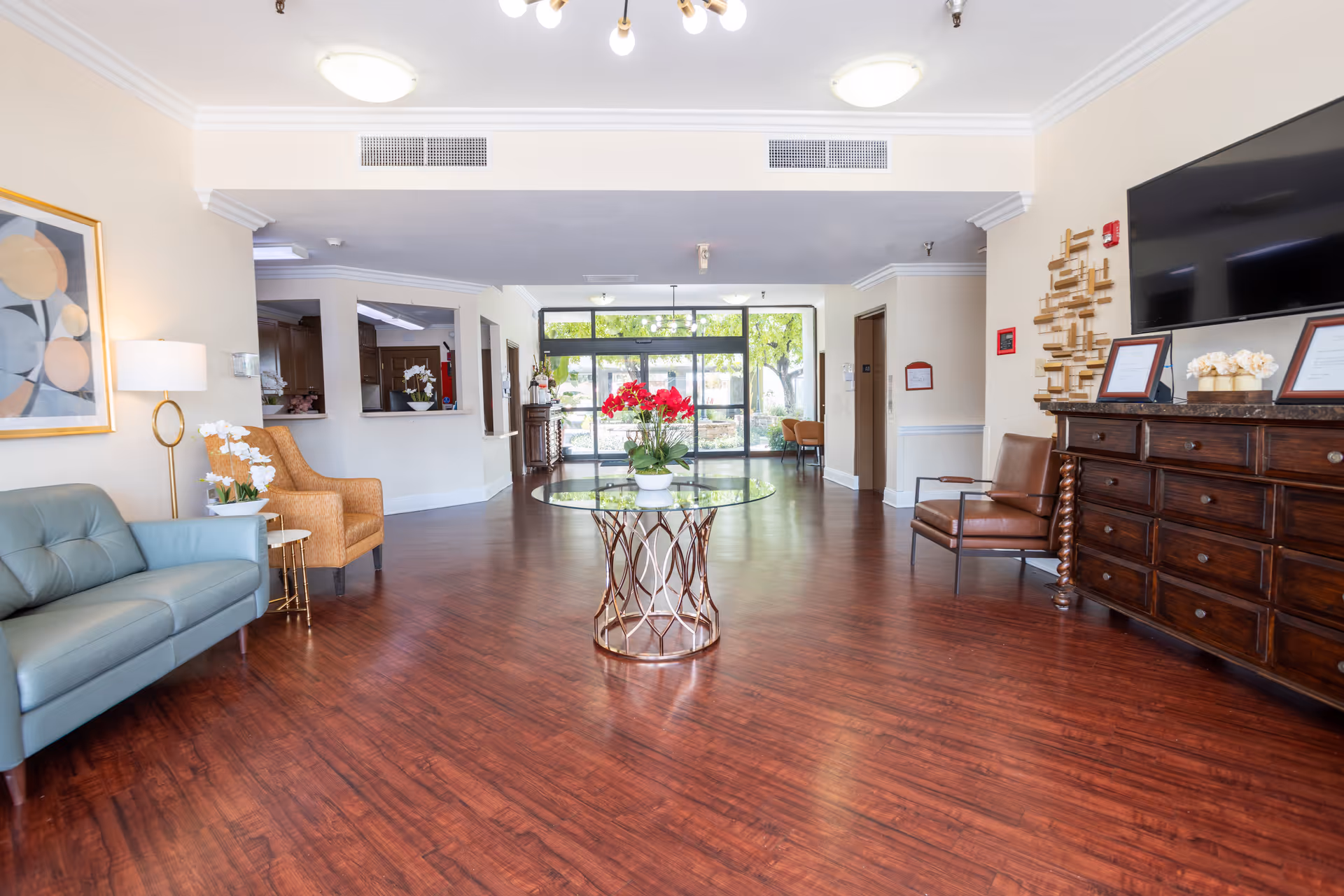 A bright and spacious senior living facility lobby with wooden flooring, a round glass table with a flower arrangement in the center, seating including a blue sofa, a tan armchair, and a brown leather chair. There is a large wooden dresser with framed certificates and a TV mounted above it on the right wall. The back of the room features large glass doors leading outside, allowing natural light to fill the space.