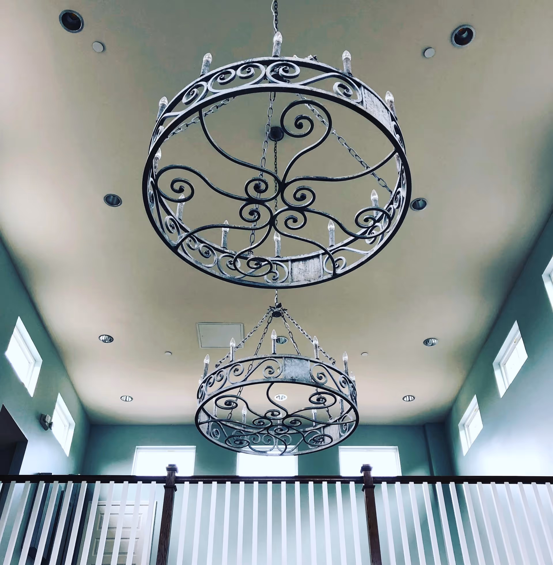 View of two large, ornate wrought iron chandeliers hanging from a high ceiling in a room with multiple small square windows near the ceiling and a white railing with wooden posts below.