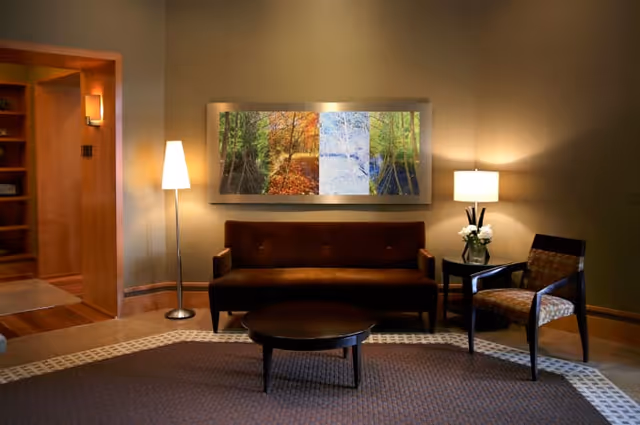 A cozy living room area with a brown sofa, a patterned armchair, a round wooden coffee table, and two lamps providing warm lighting. A large framed artwork depicting four different seasons hangs on the wall above the sofa. The room has a carpet with a decorative border and an open doorway leading to another room with wooden shelves.
