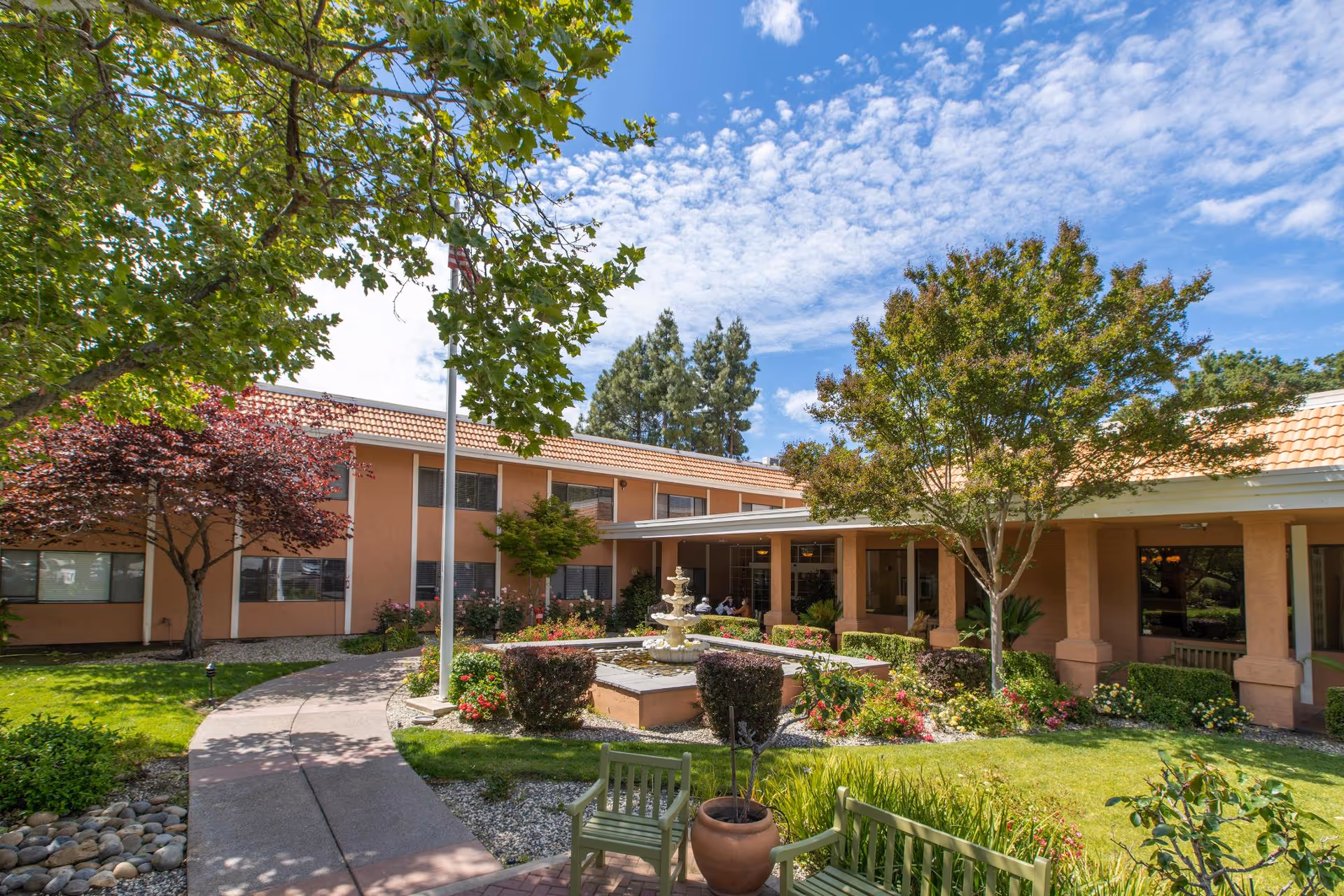 Outdoor courtyard area of a senior living facility with a paved walkway, green benches, a central water fountain, and various trees and shrubs under a partly cloudy sky.