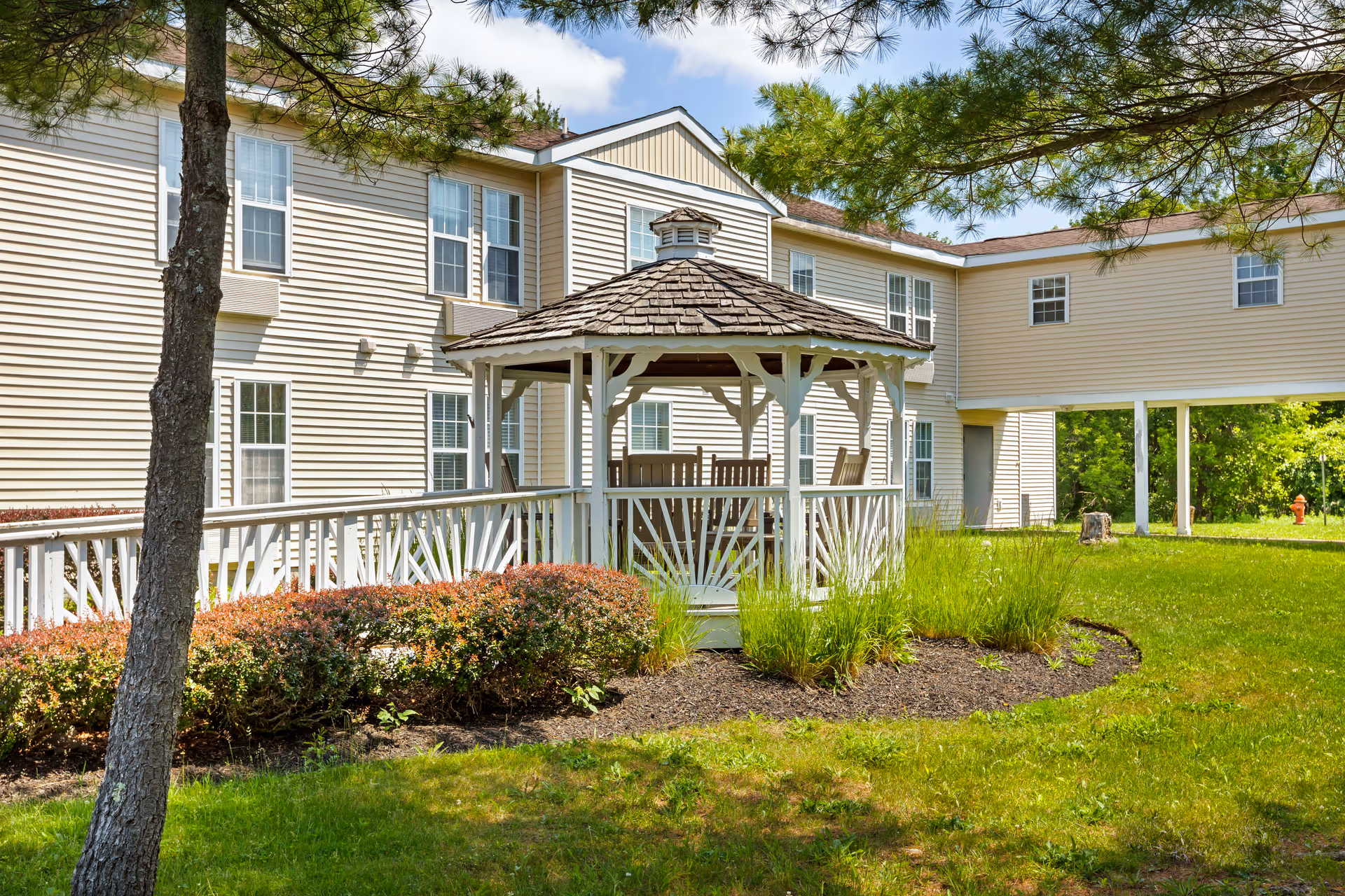 Outdoor view of a senior living facility with a white gazebo surrounded by green grass, bushes, and trees. The beige building with multiple windows is visible in the background under a partly cloudy sky.