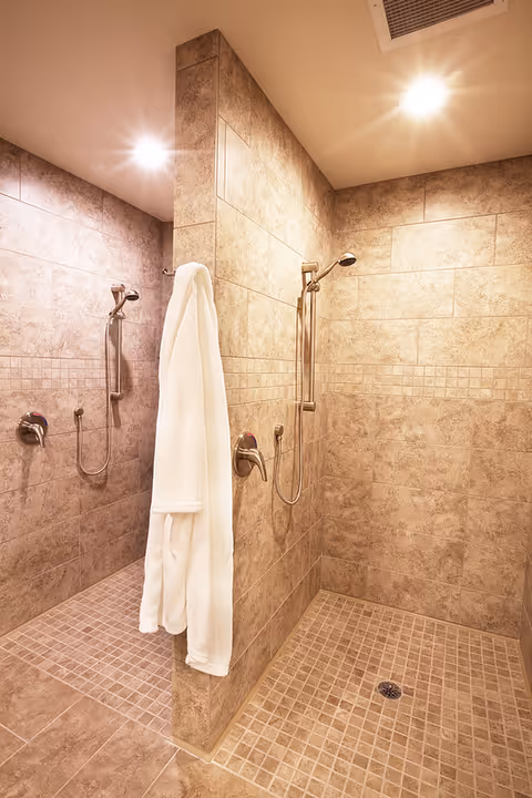 A tiled walk-in shower area with two separate shower heads and controls, separated by a tiled wall. A white bathrobe is hanging on a hook on the dividing wall. The lighting is warm and bright.