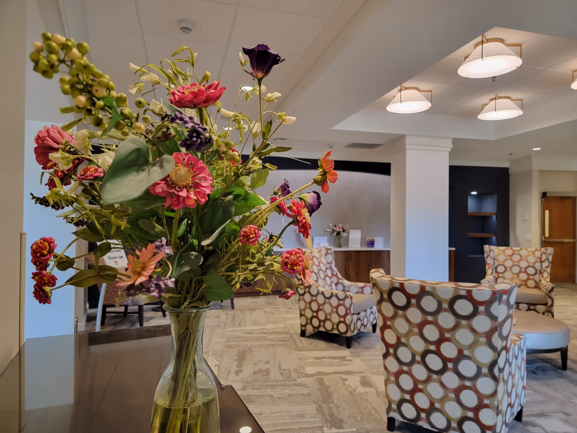 A vase with a colorful bouquet of flowers is placed on a table in the foreground. In the background, there is a seating area with patterned armchairs and a round ottoman in a well-lit room with modern ceiling lights and neutral-colored walls and carpet.