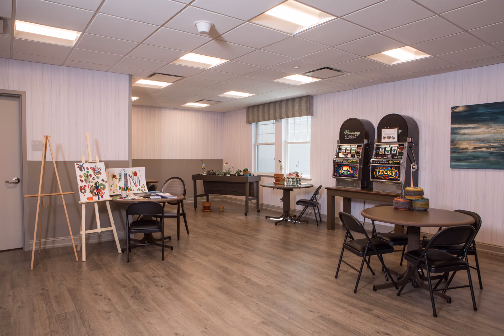 A spacious activity room with wooden flooring and soft lighting. The room features two slot machines on a table, several round tables with black folding chairs, an easel with colorful paintings, and a foosball table near the windows. The walls are light-colored with a subtle pattern, and there is a large abstract painting on one wall.