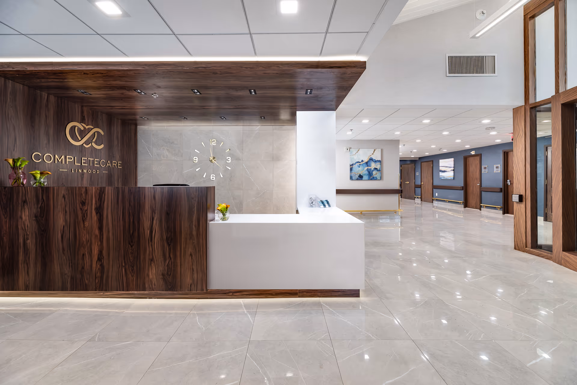 Reception area of Complete Care at Linwood featuring a modern wooden and white reception desk with small flower vases, a wall clock, and a hallway with doors and artwork on the walls.
