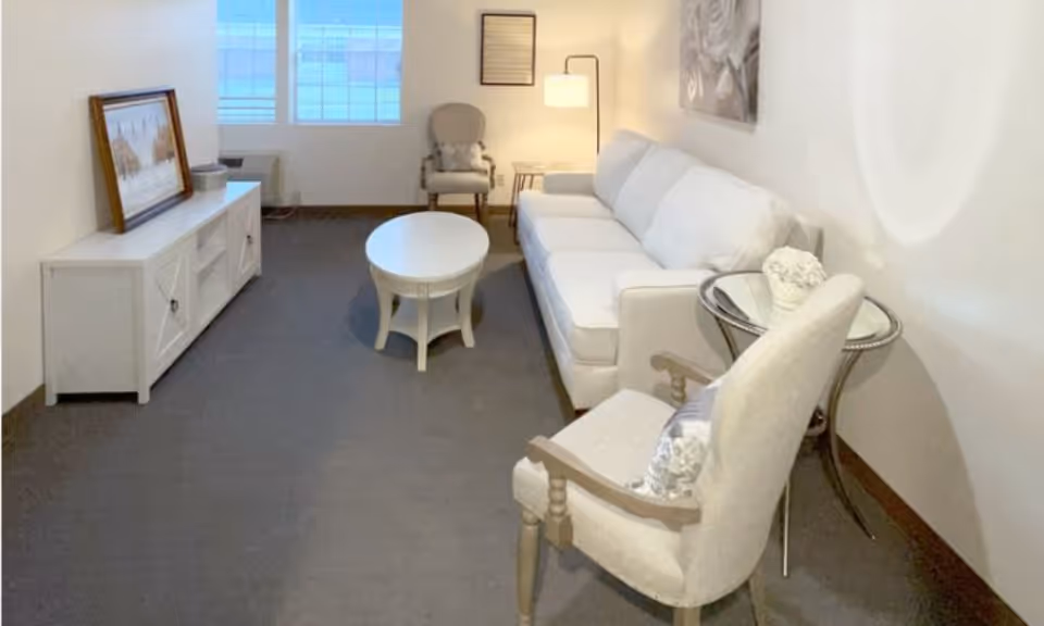 Small living room with a white sofa, two upholstered armchairs, a round coffee table, a TV console and a floor lamp under a window.