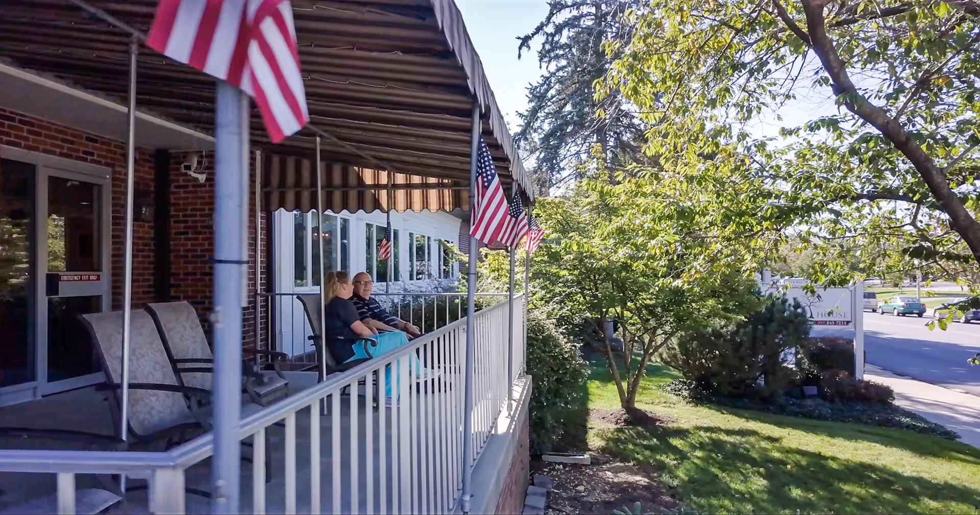 Two people sitting on a covered front porch with chairs and American flags overlooking a grassy yard and sidewalk.