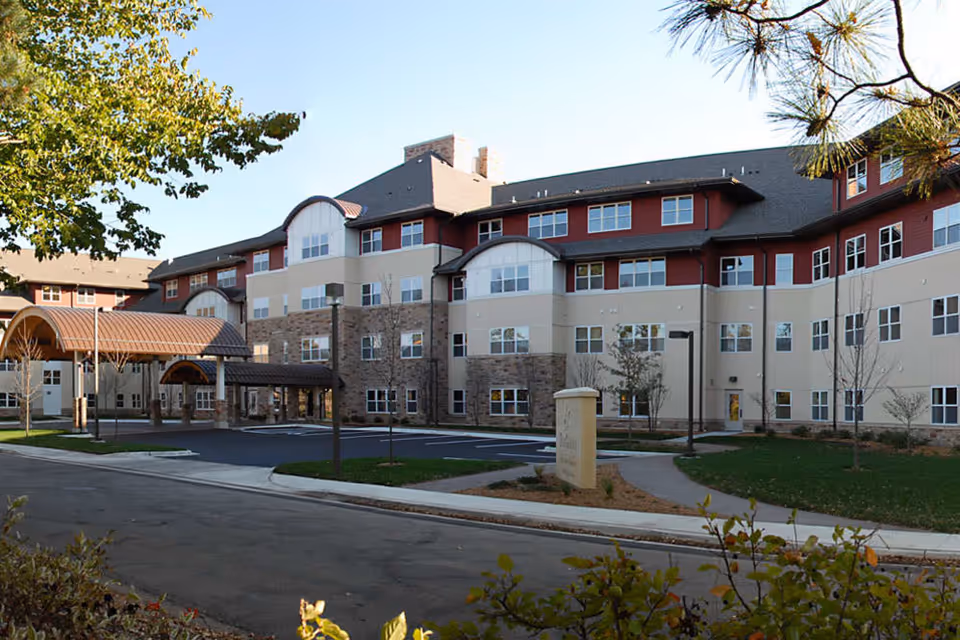 Exterior front view of a multi-story senior living building with a covered entrance, parking area, and landscaped grounds.