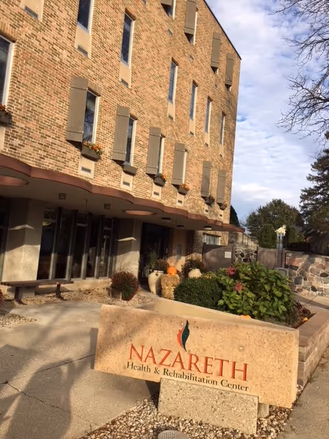 Exterior view of Nazareth Health and Rehabilitation Center showing a multi-story brick building with several windows, flower boxes, and an entrance area. In front of the building is a stone sign with the facility's name and logo. There are plants and a stone wall near the entrance under a partly cloudy sky.