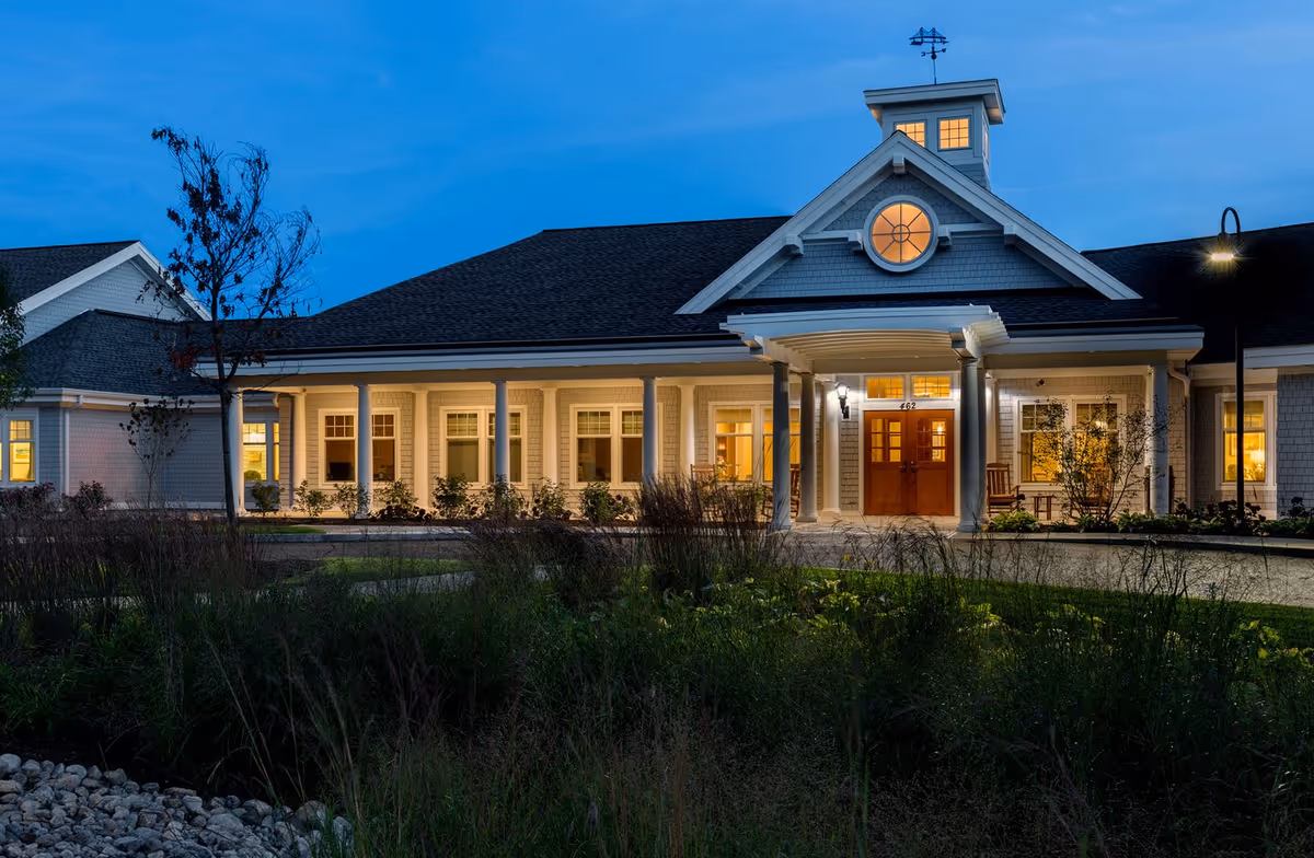 Exterior view of Bridges by EPOCH at Mashpee building at dusk, showing a well-lit entrance with columns, multiple windows, and a landscaped foreground with grass and plants.
