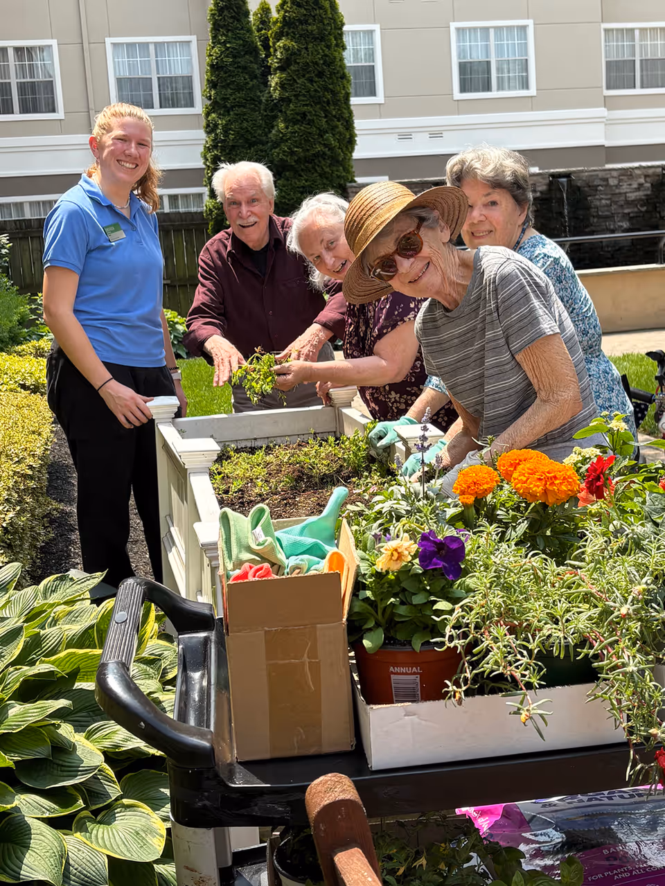 A group of four elderly people and a caregiver are gardening together outdoors at a senior living facility. They are smiling and tending to plants in raised garden beds, with various flowers and gardening tools visible. The background shows part of the building and greenery.