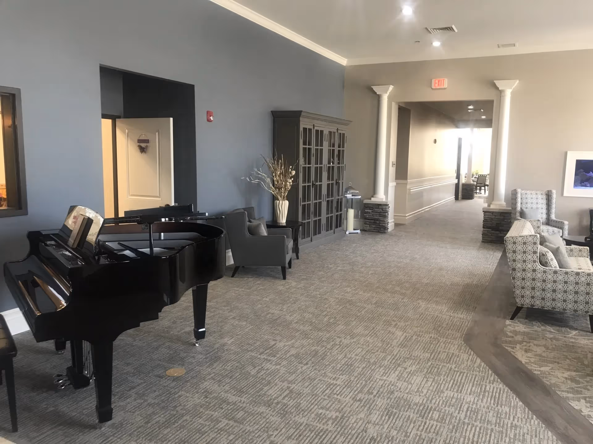 Interior view of a senior living facility lounge area featuring a black grand piano on the left, a gray armchair, a tall cabinet with glass doors, and a hallway with columns and an exit sign. On the right, there are patterned armchairs and a sofa arranged on a carpeted floor.