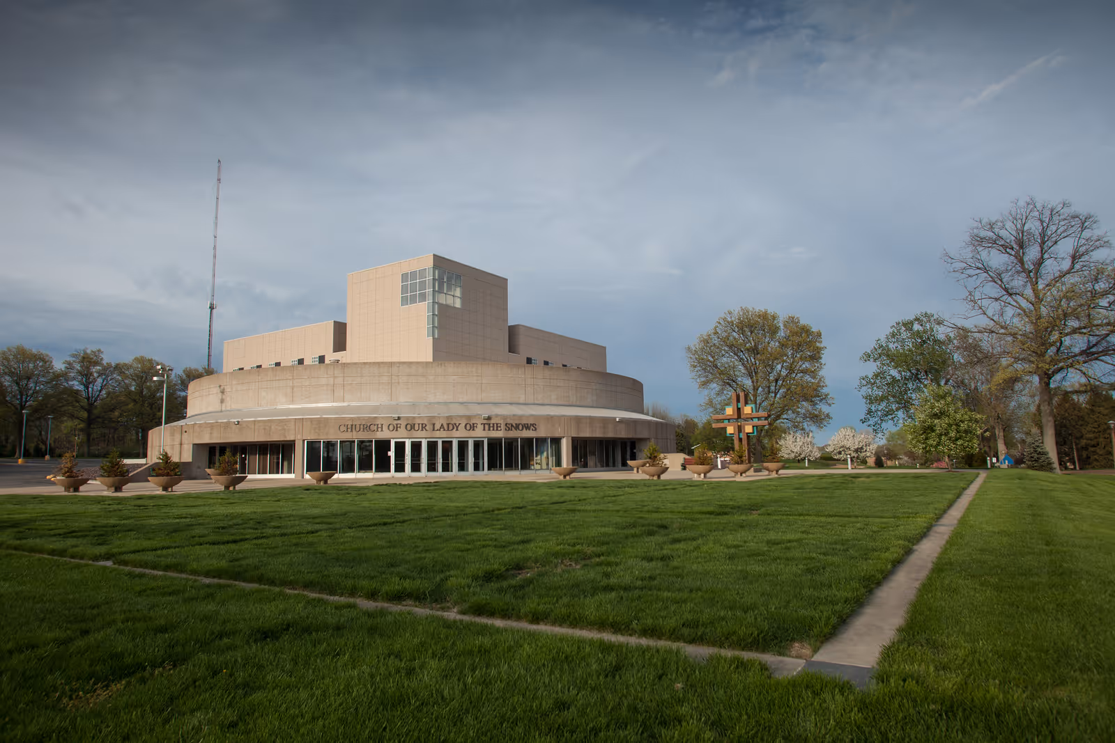 Front exterior of the Church of Our Lady of the Snows, a circular modern building with a large green lawn and a cross in front.