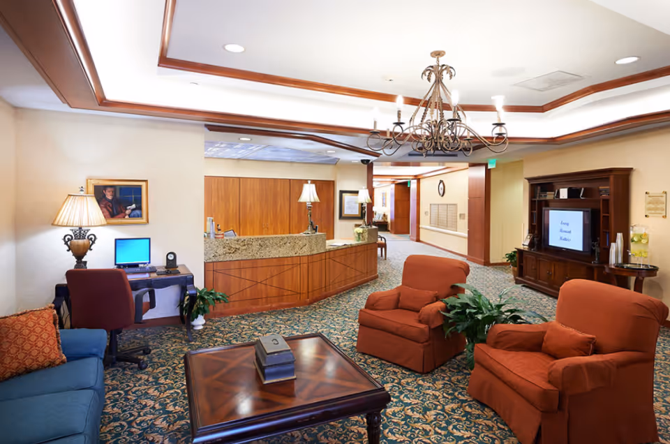 Well-lit senior living facility common area with a reception desk, seating area of armchairs and a coffee table, a TV cabinet, and a computer workstation.