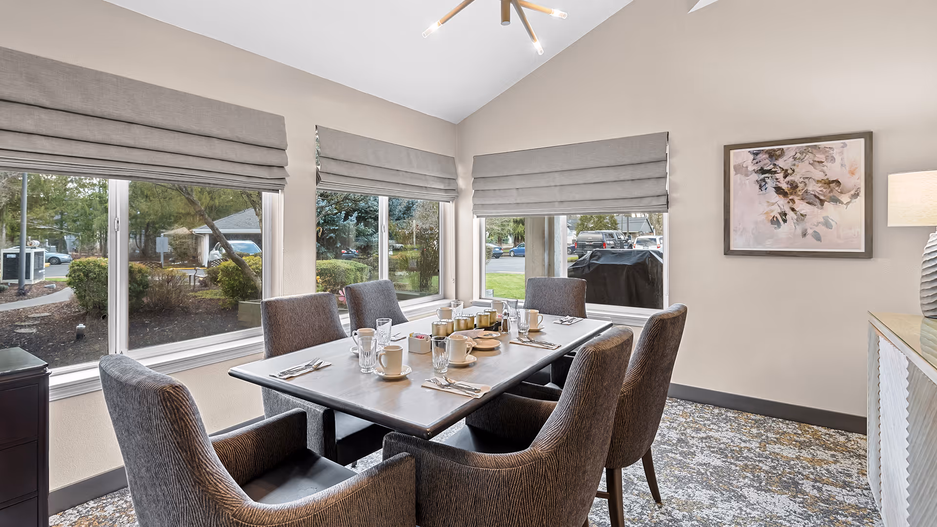 A dining room with a rectangular table set for six people. The table has cups, glasses, utensils, and condiments. There are six upholstered chairs around the table. The room has large windows with gray roman shades, a patterned carpet, a sideboard with a lamp, and a framed abstract painting on the wall. Outside the windows, greenery and parked cars are visible.