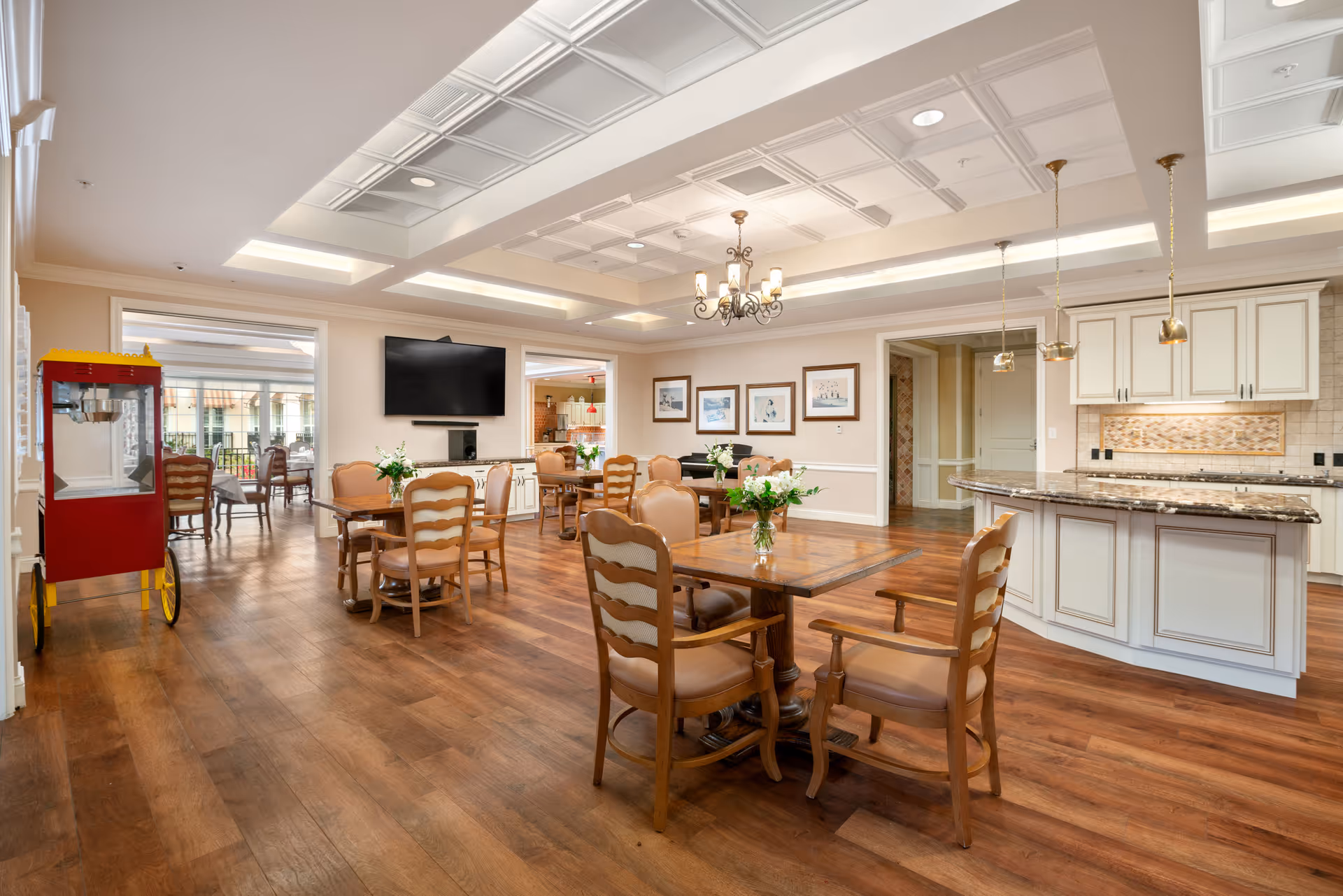 Bright communal dining area with wooden tables and chairs, a kitchen island with pendant lights, a wall-mounted TV, and a red popcorn cart.