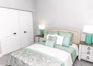A neatly made bed with a tufted beige headboard, adorned with multiple pillows in shades of white, mint green, and gray. On each side of the bed are matching white nightstands with drawers, each topped with a lamp featuring a white shade and a mint green base. To the left of the bed is a closed double-door closet with black handles. The walls are painted white, and the room has a calm, clean, and cozy atmosphere.