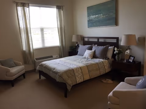A neatly made bed in a furnished bedroom with two armchairs, nightstands and lamps, a window with curtains, and wall art above the headboard.