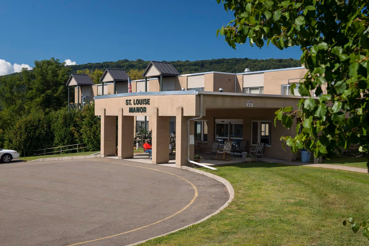 Exterior view of St. Louise Manor, a beige brick building with a covered entrance and a circular driveway. There is a person sitting on a bench under the entrance canopy. The building is surrounded by green grass, trees, and hills in the background under a clear blue sky.