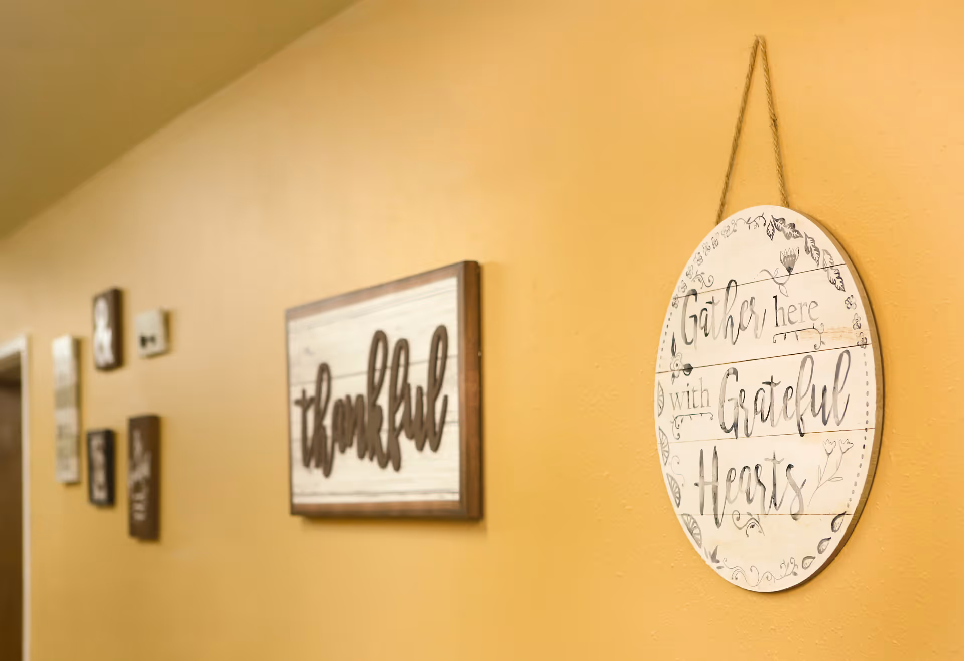 Yellow interior hallway wall decorated with wooden signs including one that says 'thankful' and a round sign reading 'Gather here with grateful hearts'.