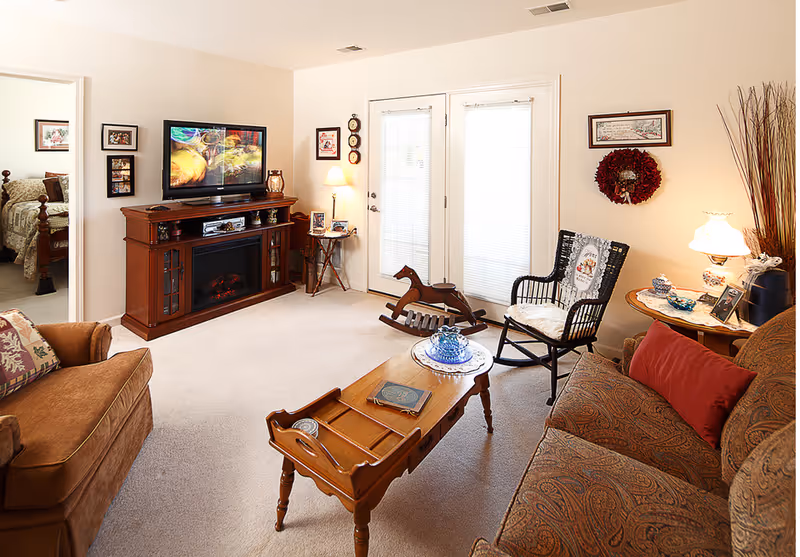 A cozy living room with beige walls and carpet. The room features a brown patterned sofa with a red pillow, a brown armchair, a wooden coffee table with decorative items, a black rocking chair with a cushion, and a wooden rocking horse near glass double doors with blinds. A wooden entertainment center with a flat-screen TV and an electric fireplace is against one wall. A small side table with a lamp and framed photos is next to the doors. The room is warmly lit and decorated with framed pictures and a red wreath on the wall.