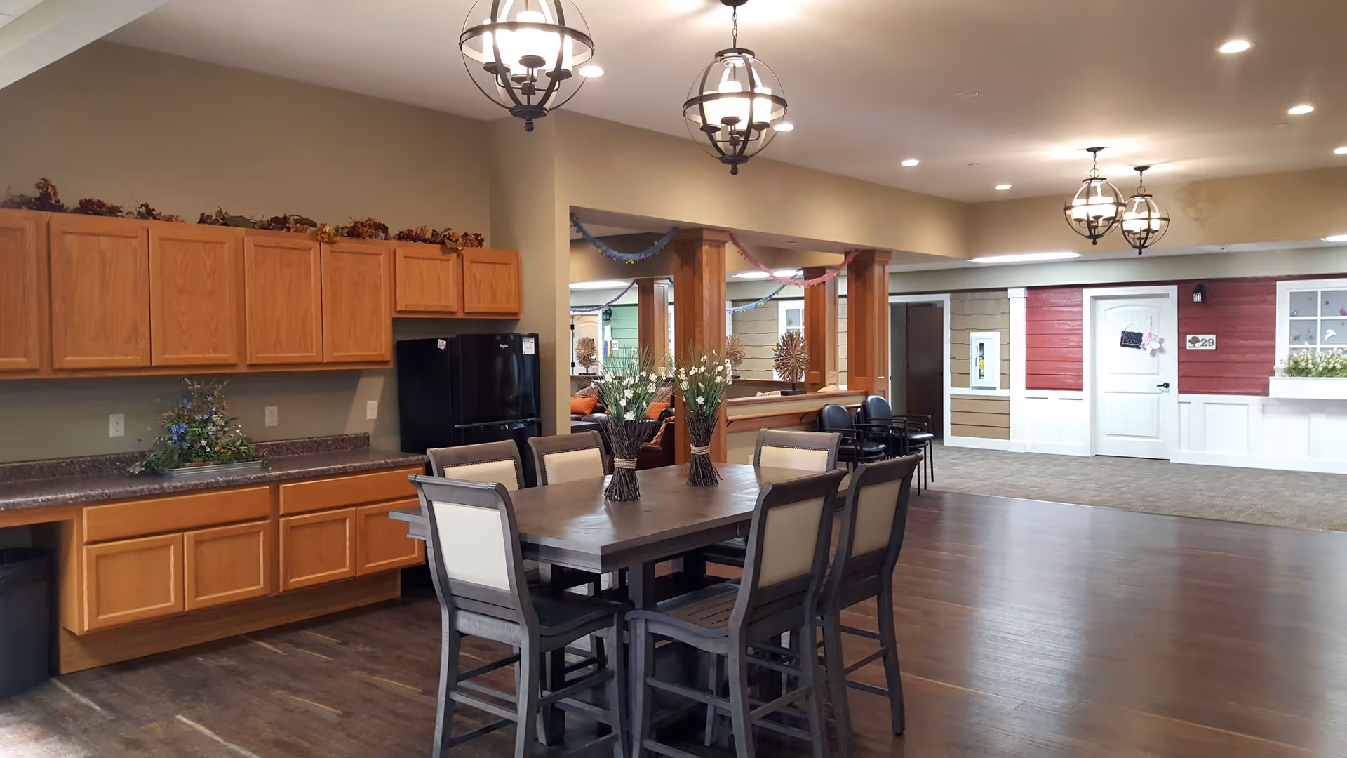 Communal dining area with a large table and chairs, kitchenette cabinets and refrigerator, and decorative pendant lights.